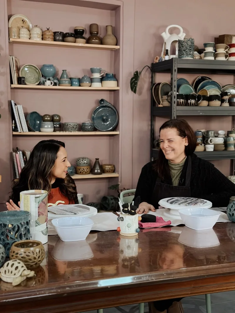 A Cristina e uma convidada à mesa comprida do ateliê, com as prateleiras de peças atrás