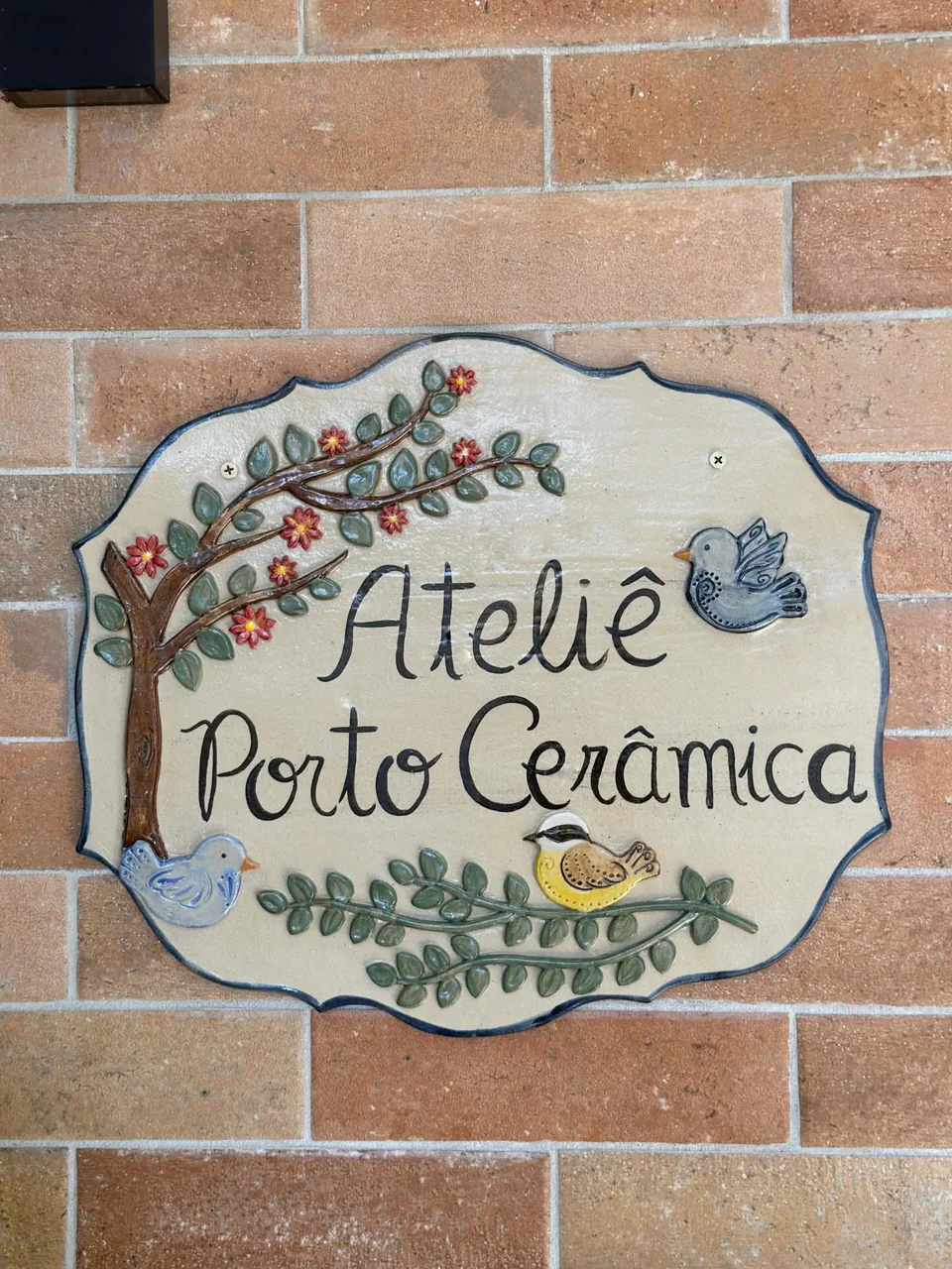 Placa de cerâmica pintada à mão na parede de tijolos, com os dizeres Ateliê Porto Cerâmica, um passarinho e flores
