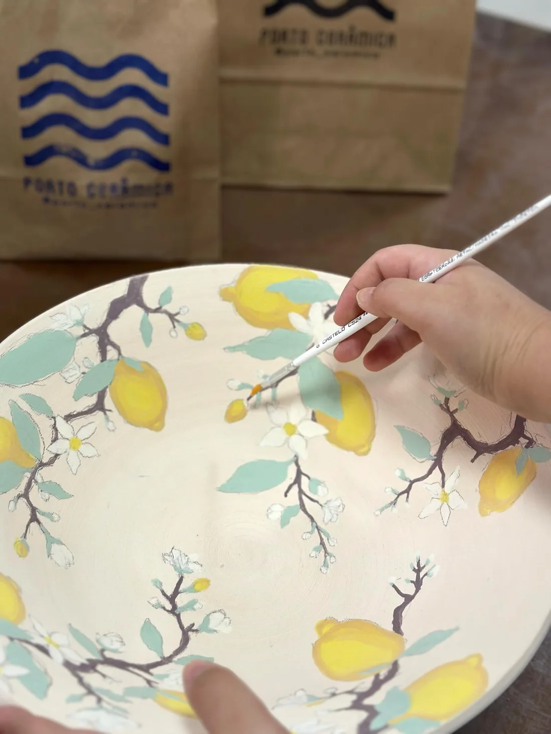 Mão pintando limões e folhas num prato de cerâmica branco, com a sacola do ateliê ao fundo