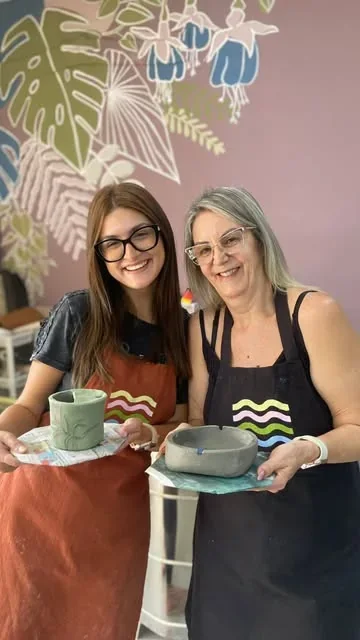 Cris e Bê de avental, sorrindo, cada uma com uma peça de cerâmica nas mãos